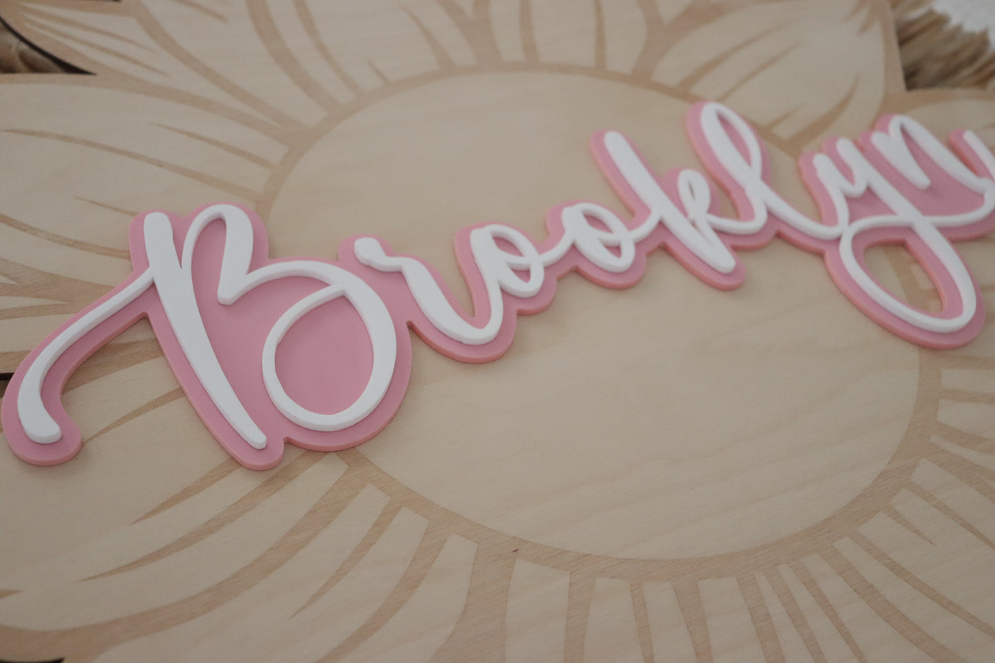A close-up of a decorative script sign reading "Brooklyn" in pink and white, mounted on a light wood sunflower plaque.
