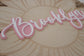 A close-up of a decorative script sign reading "Brooklyn" in pink and white, mounted on a light wood sunflower plaque.