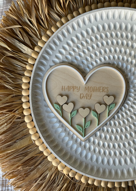 Heart-shaped wooden plaque with 'Happy Mother's Day' on a textured white surface with raffia border.